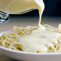 vegan cream sauce poured from glass gravy bowl onto plate of pasta